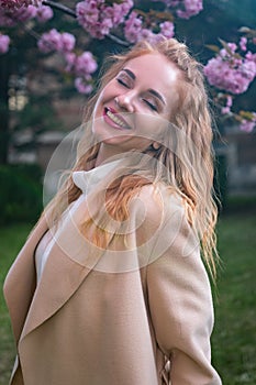 Beautiful gentle girl with wavy red hair in beige coat in blooming park enjoying spring day. Vertical frame