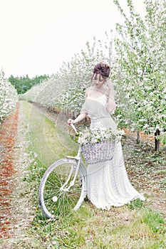 Beautiful gentle lovely girl bride in a white dress standing with his bicycle in blossoming apple orchard