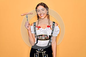 Beautiful german woman with blue eyes wearing oktoberfest dress holding fork with sausage with a happy face standing and smiling