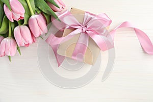 Beautiful gift box with bow and pink tulip flowers on white wooden background, flat lay. Space for text