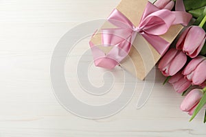 Beautiful gift box with bow and pink tulip flowers on white wooden background, flat lay. Space for text