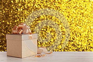 Beautiful gift box on white wooden table against shiny golden background, bokeh effect. Space for text