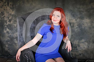 Beautiful ginger-haired woman in blue dress sitting in gray armchair