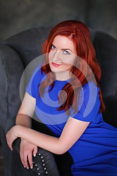 Beautiful ginger-haired woman in blue dress sitting in gray armchair