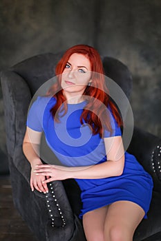 Beautiful ginger-haired woman in blue dress sitting in gray armchair