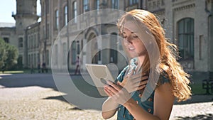 Beautiful ginger young woman typing on tablet, wind ruffling her hair, standing on street and smiling, happy and