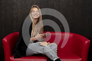 A beautiful girl of 20-25 years old holds a phone in her hands while sitting on a red sofa.