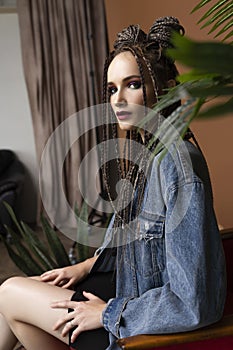 Beautiful girl with an afro cornrows hairstyle, wearing a casual denim jacket, is resting on a chair standing on an old carpet