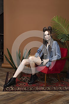 Beautiful girl with an afro cornrows hairstyle, wearing a casual denim jacket, is resting on a chair standing on an old carpet