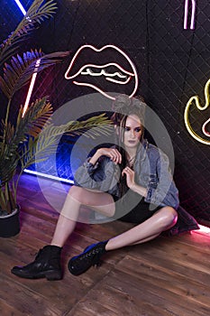 Beautiful girl with a afro cornrows, wearing denim jacket, sits on the floor in a night club at the wall made of rabitz net,