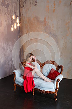 Beautiful girl in an amazing red dress on a background of minimal rustic loft wall sitting on white retro leather sofa