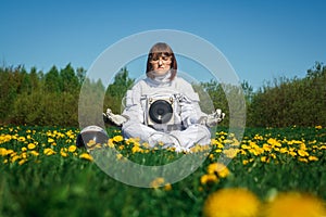 Beautiful girl astronaut without a helmet sits on a green lawn among flowers in a meditative position