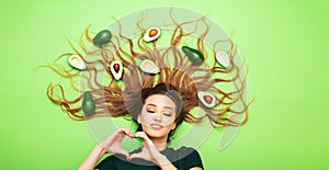 Beautiful girl with avocado fruits on long hair, young woman shows shape of heart with hands, concept love taste dieting