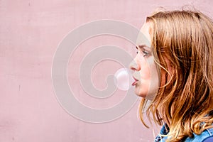 A beautiful girl on an background of a pink tint stands in profile and puffs a ball of chewing gum.