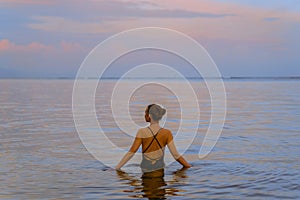 A beautiful girl bathes in the warm sea at sunset. The calm sea reflects the sunset colors of the sky.