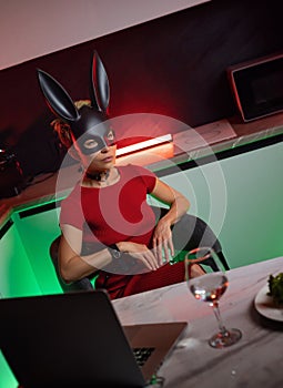 A beautiful girl in a bdsm-style rabbit mask and a bright red dress with leather straps is posing sweetly in the kitchen