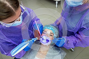 Beautiful girl in a bib looks patient& x27;s teeth and orange UV glasses in a dental clinic.