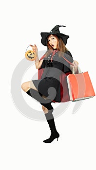 Beautiful girl in black dress and witch hat is holding pumpkin with shopping bags and smiling on white background for Halloween