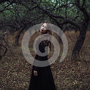 Beautiful girl in in black vintage dress with curly hair posing in the woods. Woman in retro dress lost in the forest. Worried sen