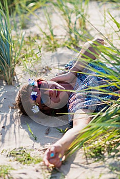 A beautiful girl in a blue dress and sunglasses lies on the sand among thick grass.