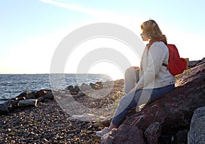 Beautiful girl body positive looking ahead on rocks seaside background in autumn season, red backpack, lifstyle