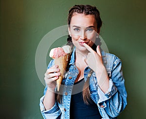 Beautiful girl with braids sitting in a cafe and eating huge multi-colored ice cream in a waffle cone. Licking finger