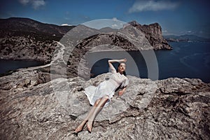 A beautiful girl, bride, in a white dress, barefoot lies on a rock against the background of a beautiful landscape, ocean and moun