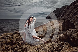 Beautiful girl, bride, in white dress, barefoot sitting on stones against the background of beautiful landscape, ocean and mountai