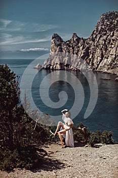 A beautiful girl, a bride, in a white dress sits a tree, on a cliff against the background of the ocean and mountains.