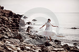 A beautiful girl, a bride, in a white dress, standing barefoot on stones, spinning her dress, against the background of the ocean.