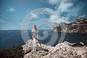 A beautiful girl, a bride, in a white dress stands on a cliff against the background of the sea and mountains.