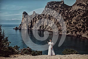 A beautiful girl, a bride, in a white dress stands on a cliff against the background of the sea and mountains.