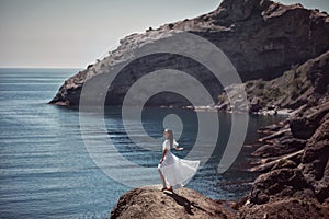 A beautiful girl, bride, in a white dress stands on a cliff against the background of the sea and mountains, her dress develops fr