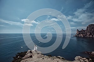 A beautiful girl, a bride, in a white dress, stands with her back on a rock against the background of the sky and ocean, birds fly