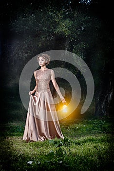 Beautiful girl with brown curly hair in wonderful dress with lamps light in fairytale forest. Art photo.