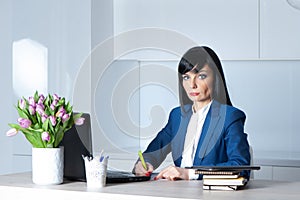 Beautiful girl, brunette, in a suit at her desk signs the papers and looks into the camera. Office work concept, businesswoman,