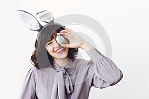 Beautiful girl in bunny ears holding easter egg near face and smiling on white background indoors, space for text. Easter hunt