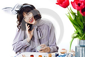 Beautiful girl in bunny ears talking on phone and decorating easter egg at table with paint, brushes, tulips in vase. Stylish