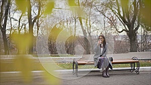 Beautiful girl in a coat sitting on a bench in a sunny park in autumn. Girl drinks warm coffee and outdoors. Portrait of