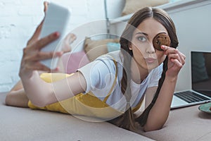 beautiful girl with cookie taking selfie on smartphone while lying on sofa