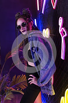 Beautiful girl with a cornrows hairstyle, wearing denim jacket and sunglasses, posing in a night club at the rabitz net wall,