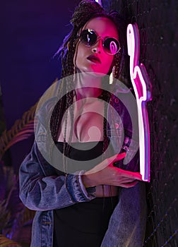 Beautiful girl with a cornrows hairstyle, wearing denim jacket and sunglasses, posing in a night club at the rabitz net wall,