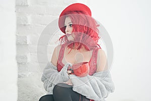 A beautiful girl in cozy warm clothes holds a large Cup of drink near the window sill at the window in her white room