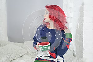 A beautiful girl in cozy warm clothes holds a large Cup of drink near the window sill at the window in her white room