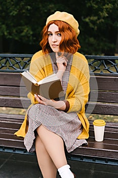 A beautiful girl with a curly hairstyle is sitting on a park bench and reading