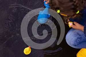 Beautiful girl in denim suit and yellow rubber boots plays with plastic duck and whale in a puddle