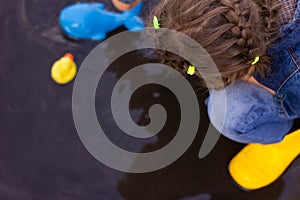 Beautiful girl in denim suit and yellow rubber boots plays with plastic duck and whale in a puddle