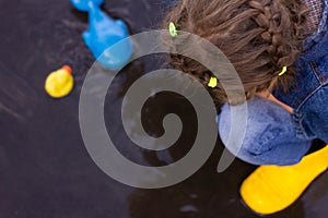 Beautiful girl in denim suit and yellow rubber boots plays with plastic duck and whale in a puddle