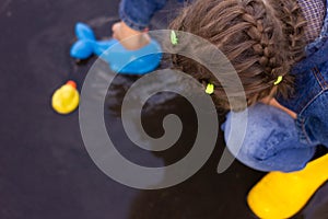 Beautiful girl in denim suit and yellow rubber boots plays with plastic duck and whale in a puddle