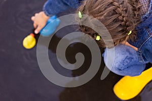 Beautiful girl in denim suit and yellow rubber boots plays with plastic duck and whale in a puddle
